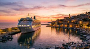 Ein Kreuzfahrtschiff liegt bei Sonnenuntergang im Hafen. Die Stadtlichter spiegeln sich im Wasser, umgeben von sanften Farben des Sonnenuntergangs.